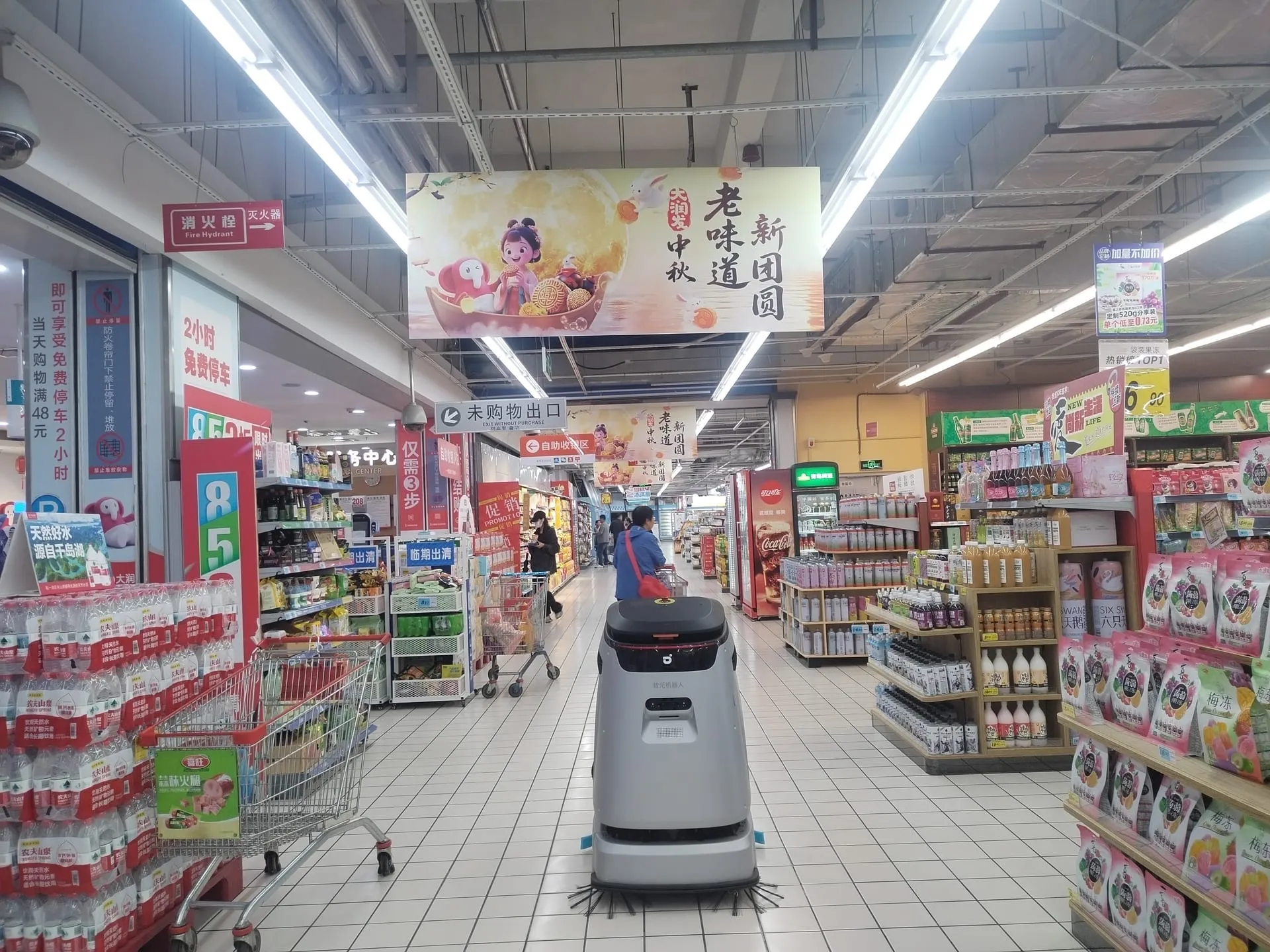 A cleaning robot working through a busy supermarket aisle with shoppers around